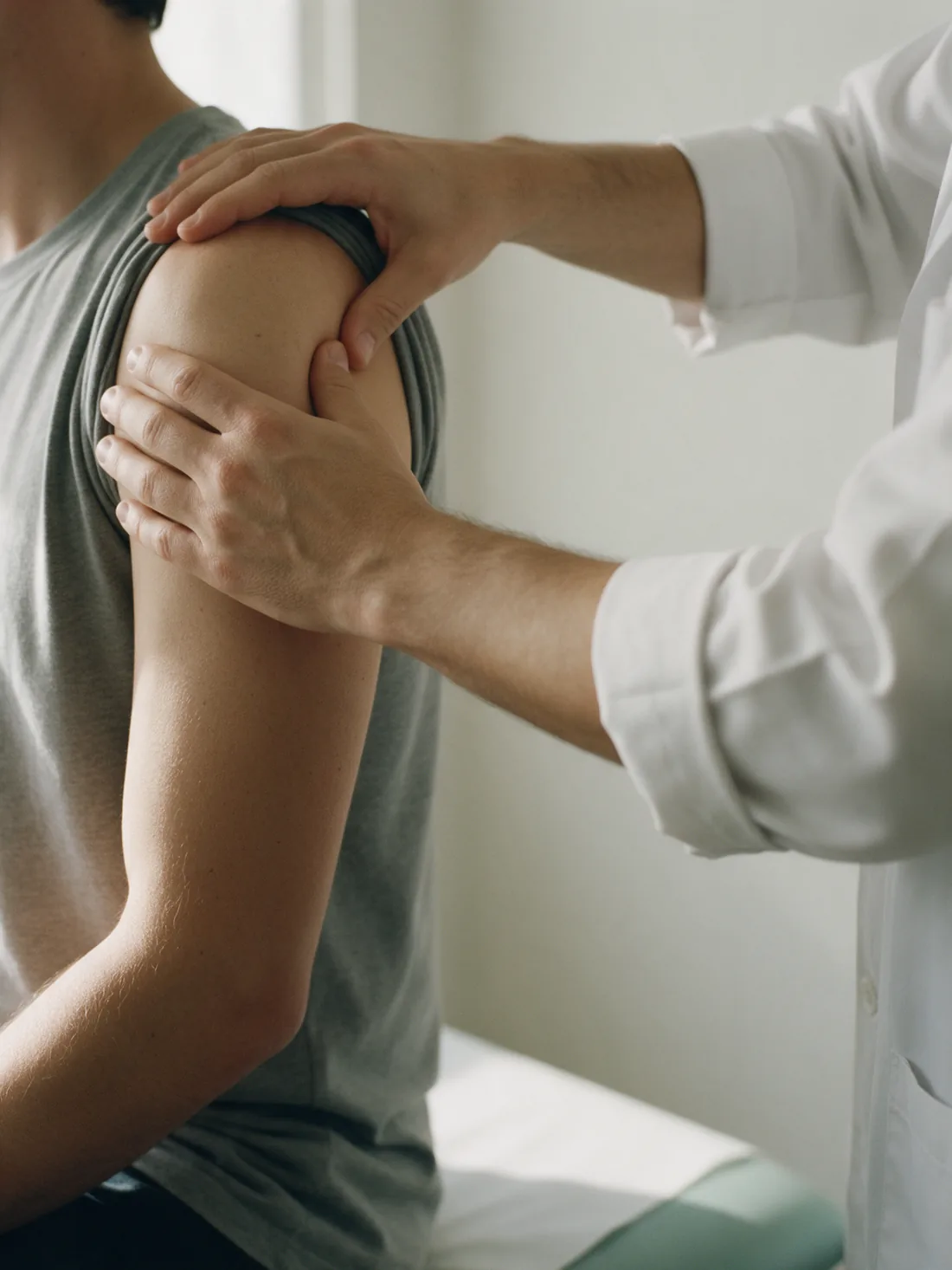 Exame físico do ombro: as duas mãos de um médico de jaleco apalpam a articulação de um paciente sentado.